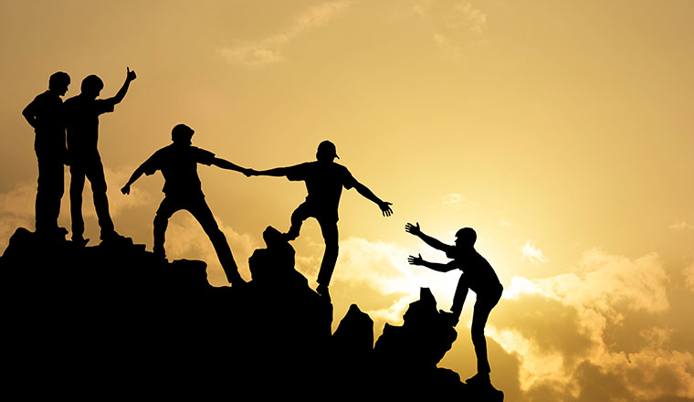 Photo of people helping each other on a golden sky background. 4 people are helping a 5th person up a mountain. 